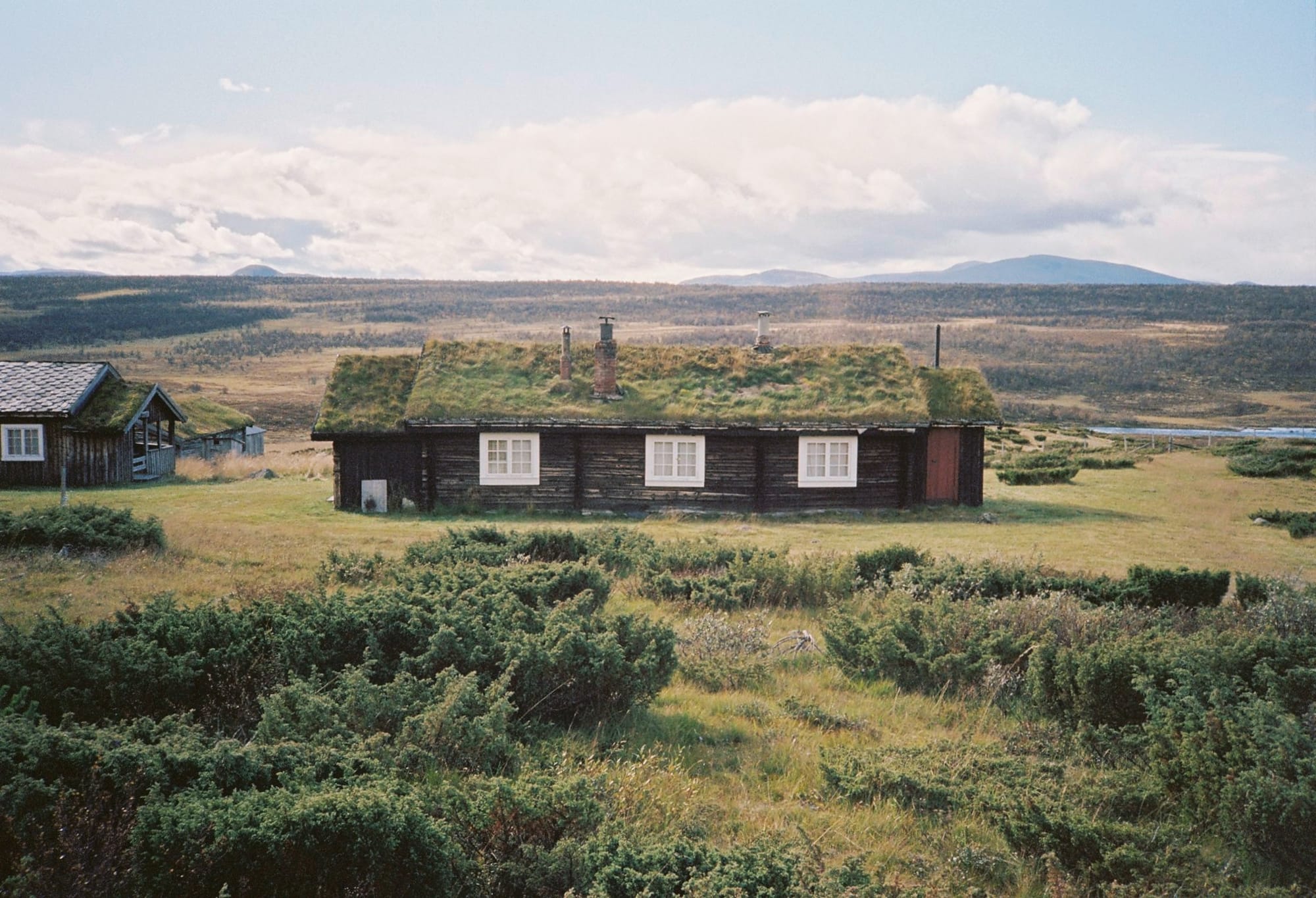 Traditional Norwegian grass-roofed wooden cabin in mountain landscape with cloudy sky and distant hills.