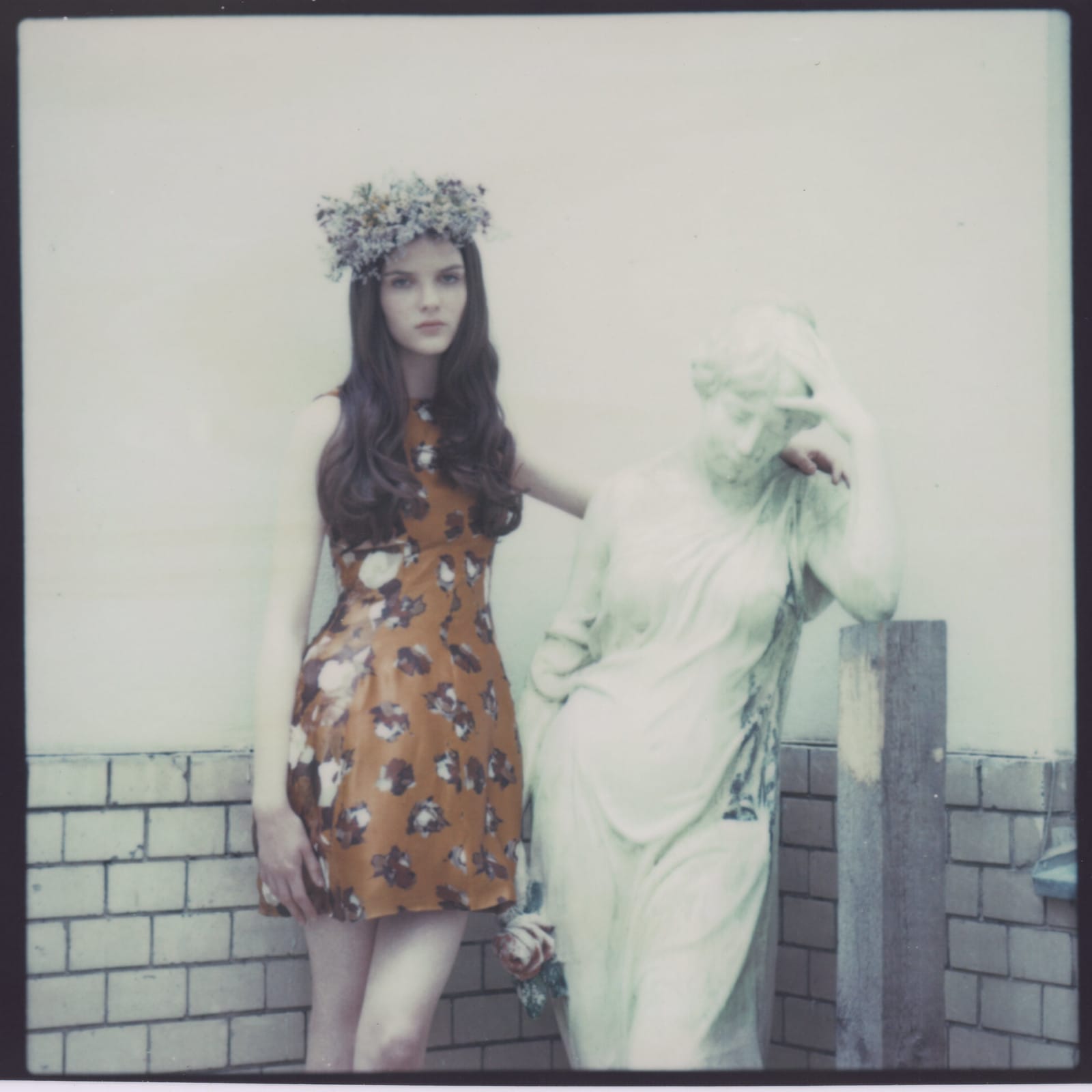 Woman in floral print dress wearing flower crown standing beside classical statue on rooftop.