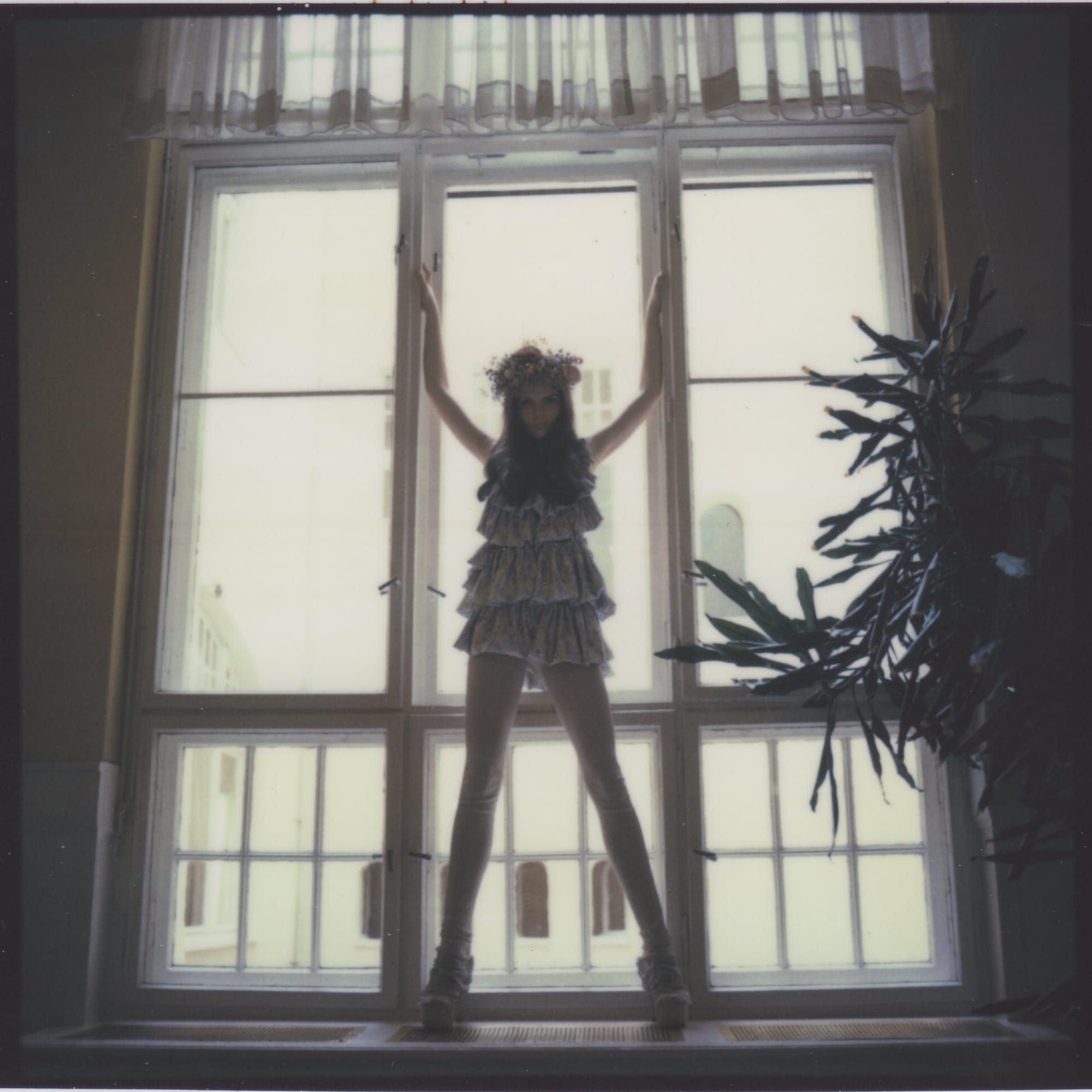 Silhouette of woman in tiered dress with flower crown standing at tall window backlit.