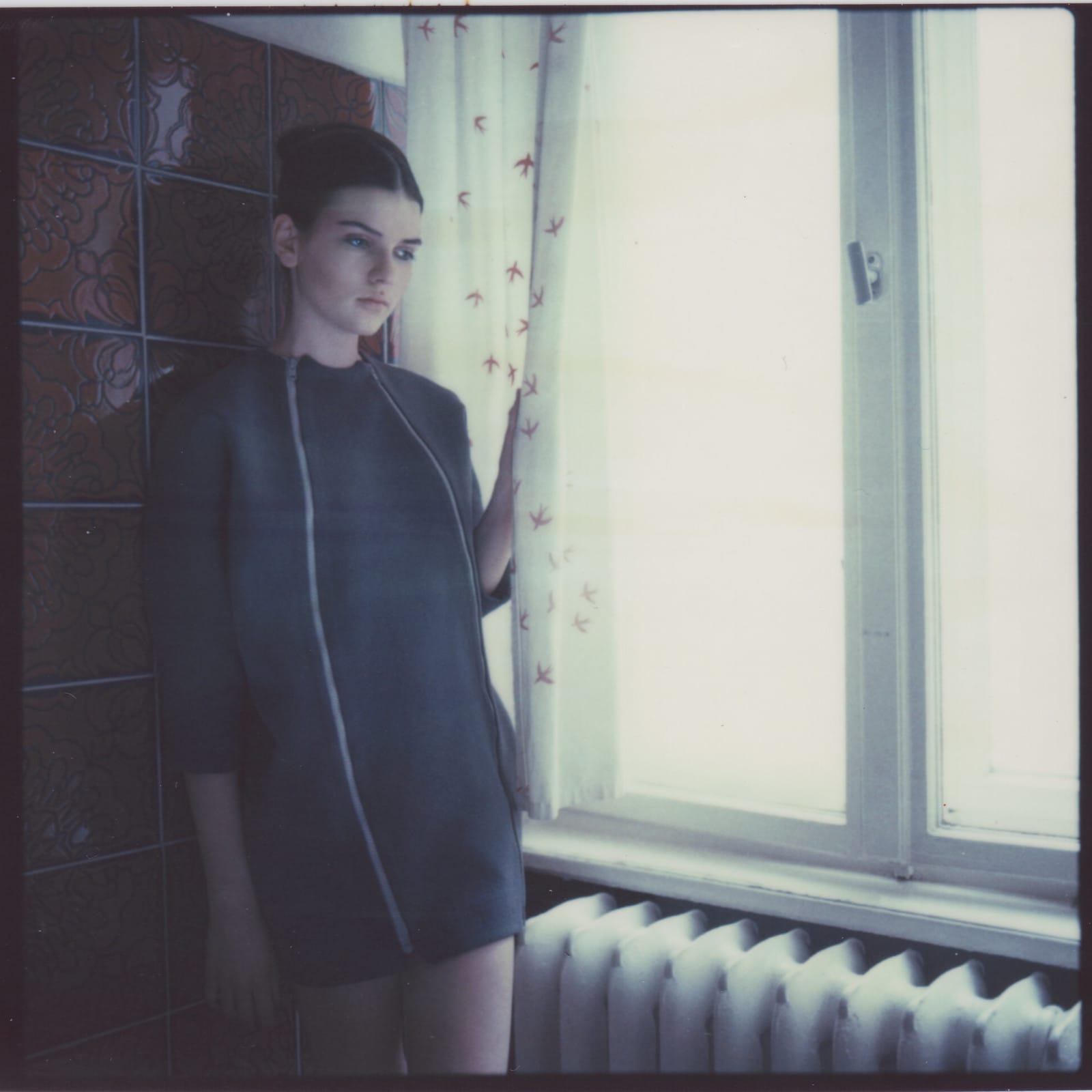 Woman in dark jacket by window with patterned curtains in pale green tinted square format film.