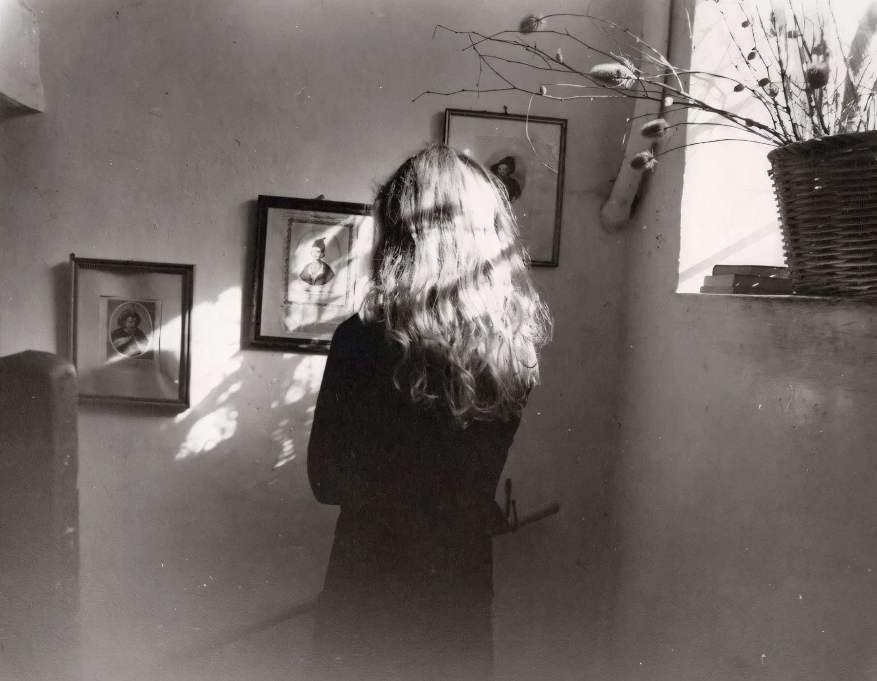 Woman with long hair photographed from behind in room with framed photos and dried branches.