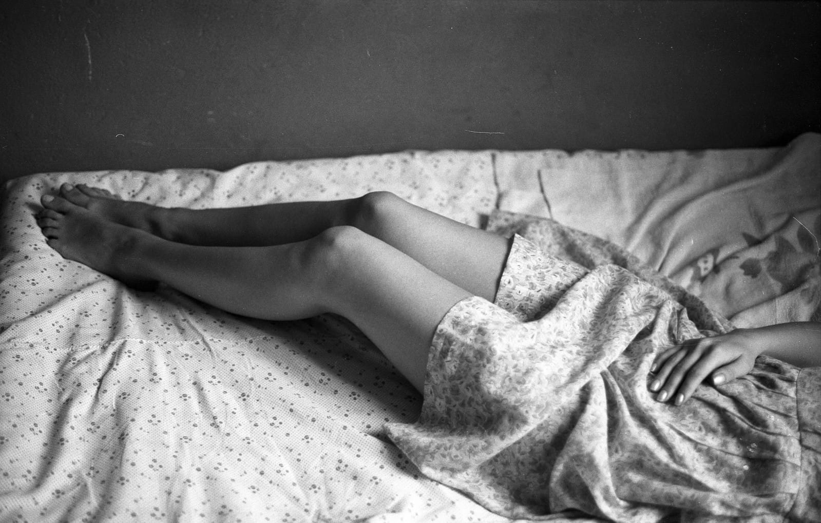 Black and white intimate photograph of bare legs and feet resting on floral patterned bed sheets.