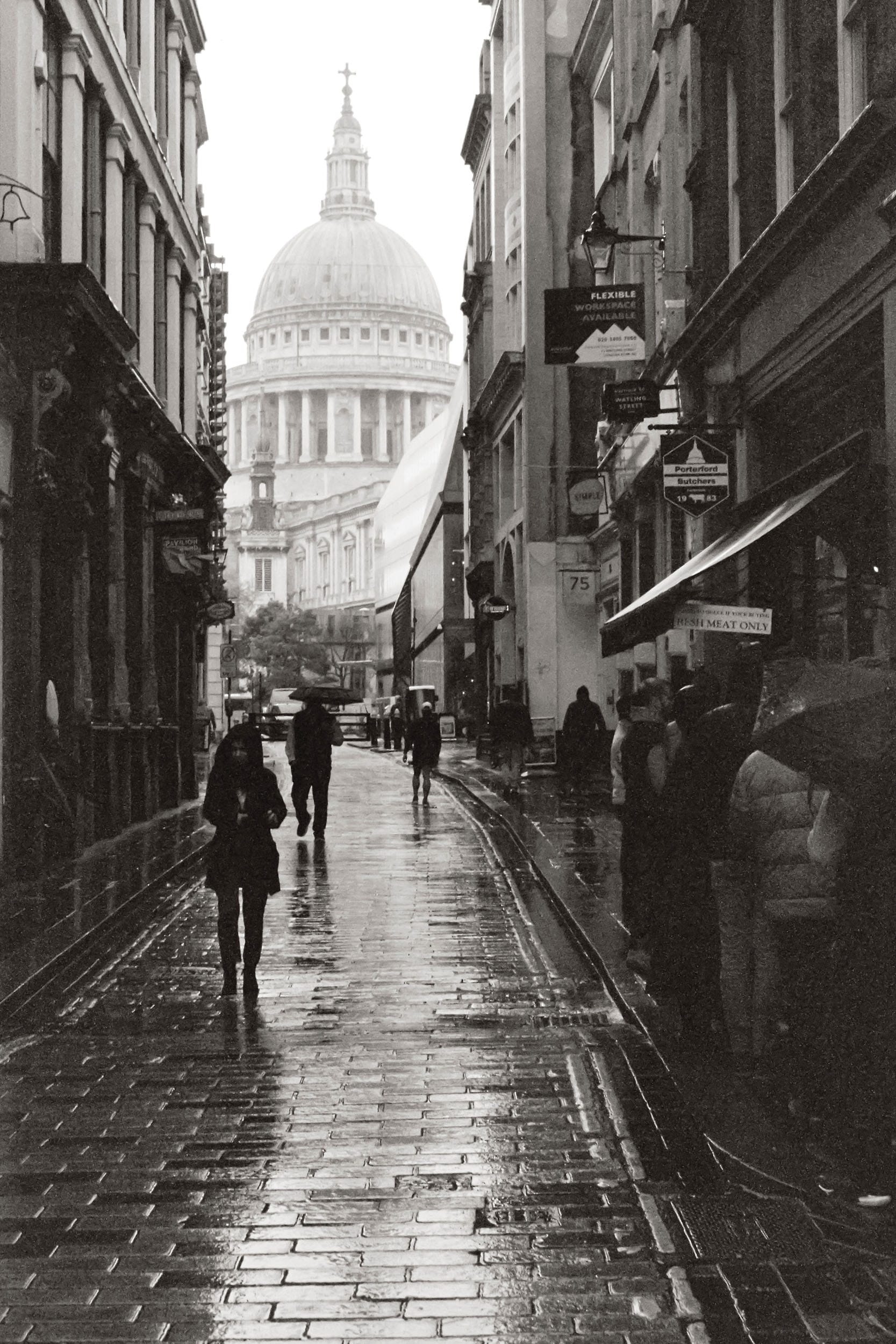 Black and white rainy London street with St Pauls Cathedral dome visible between buildings.