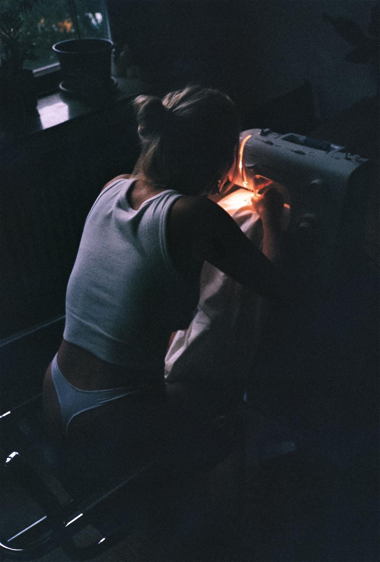 Dark moody portrait of a lingerie-clad woman sewing under a warm light.