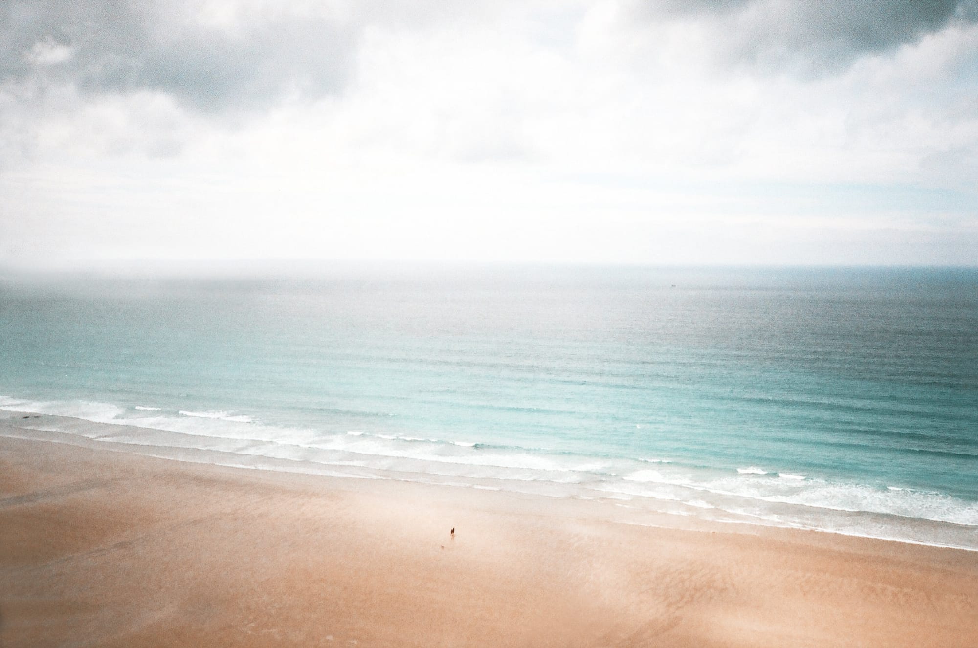 Minimalist coastal seascape with soft waves rolling onto empty sandy beach under overcast sky.