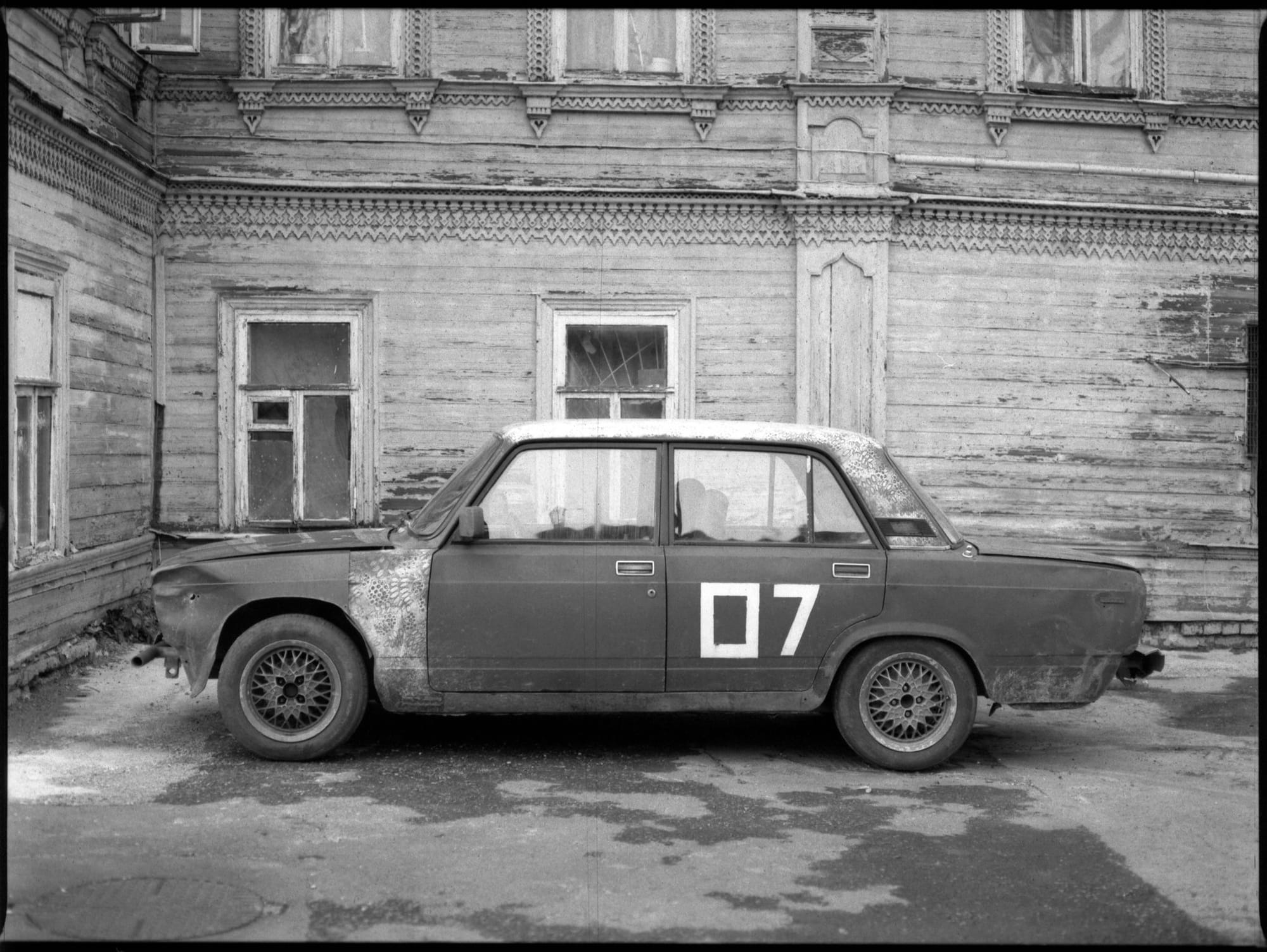 Black and white Soviet car with number 07 parked beside weathered ornate wooden building.