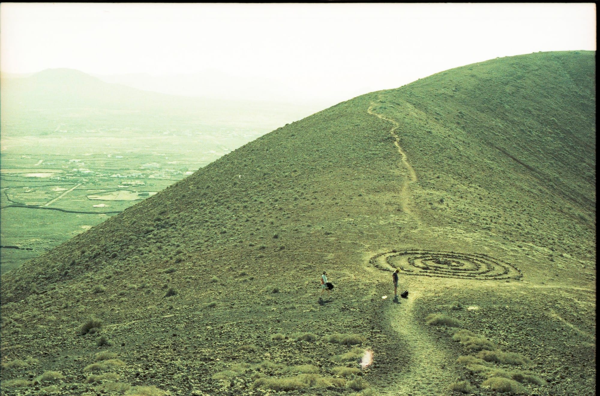 Green mountainside with spiral pattern and two small figures on hiking trail.