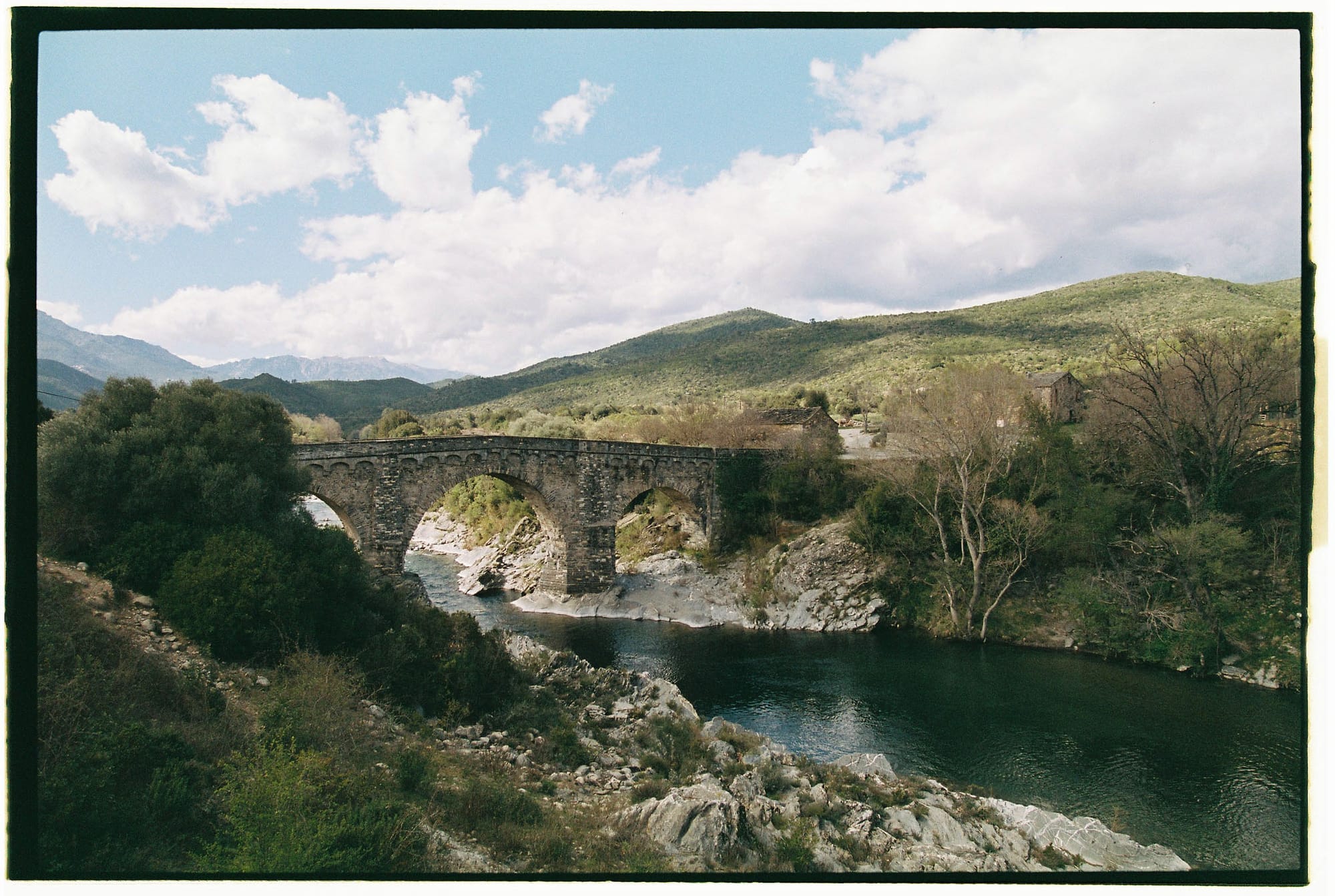 Ancient stone arch bridge over river with green hills and cloudy sky in background.
