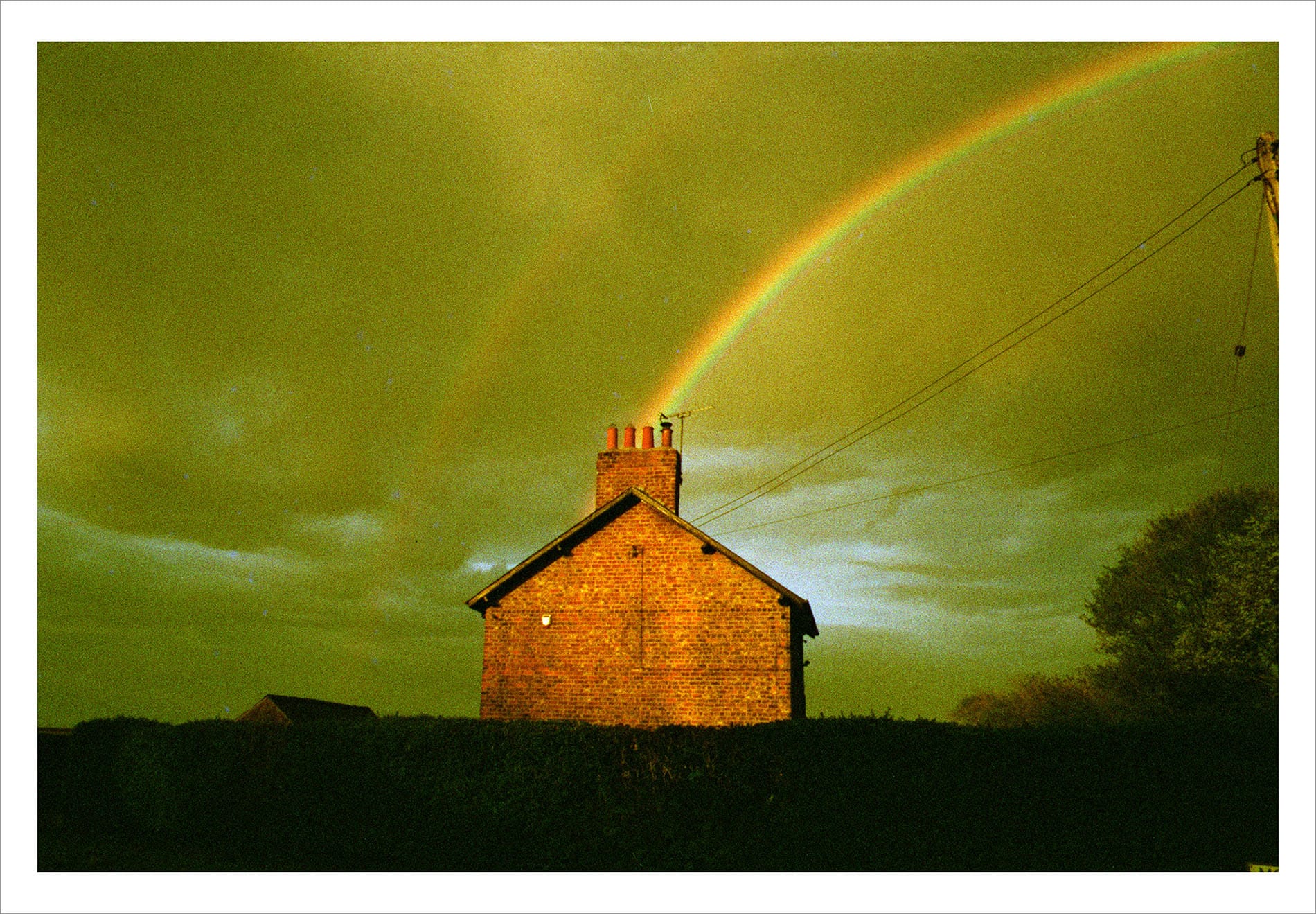 Brick house silhouette with rainbow and power lines against dramatic yellow-green stormy sky.