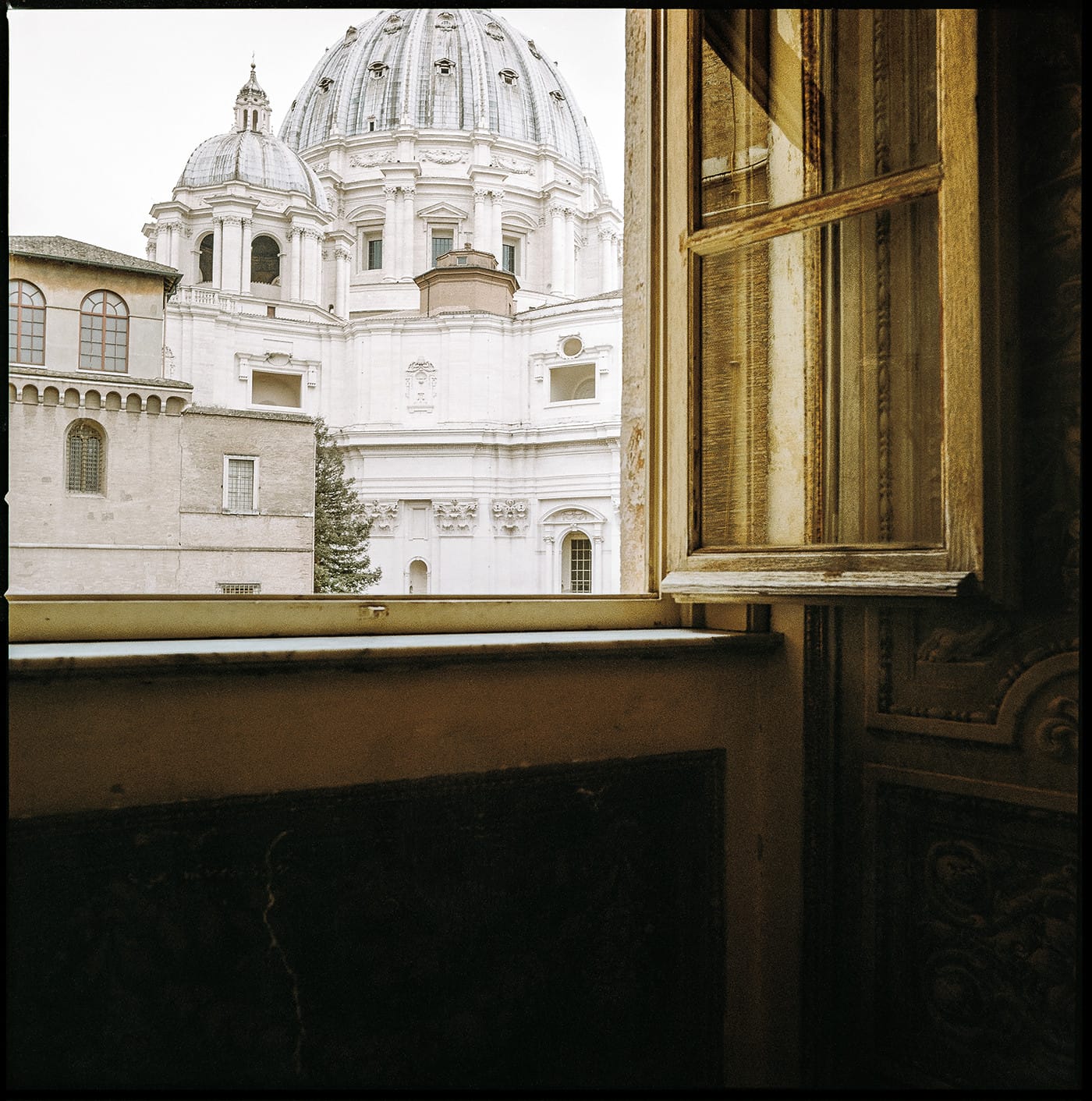 Vatican dome viewed through open window with curtains in warm interior light.