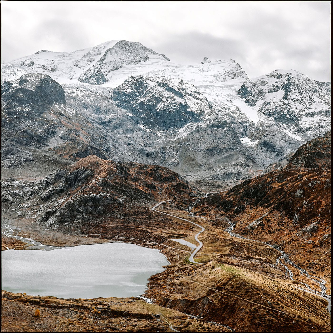 Alpine lake with winding mountain road leading through brown hillsides toward snow-covered peaks.