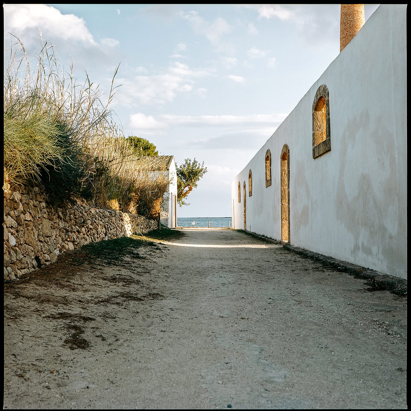 Dirt road between white wall and stone wall leading toward ocean view Greece.