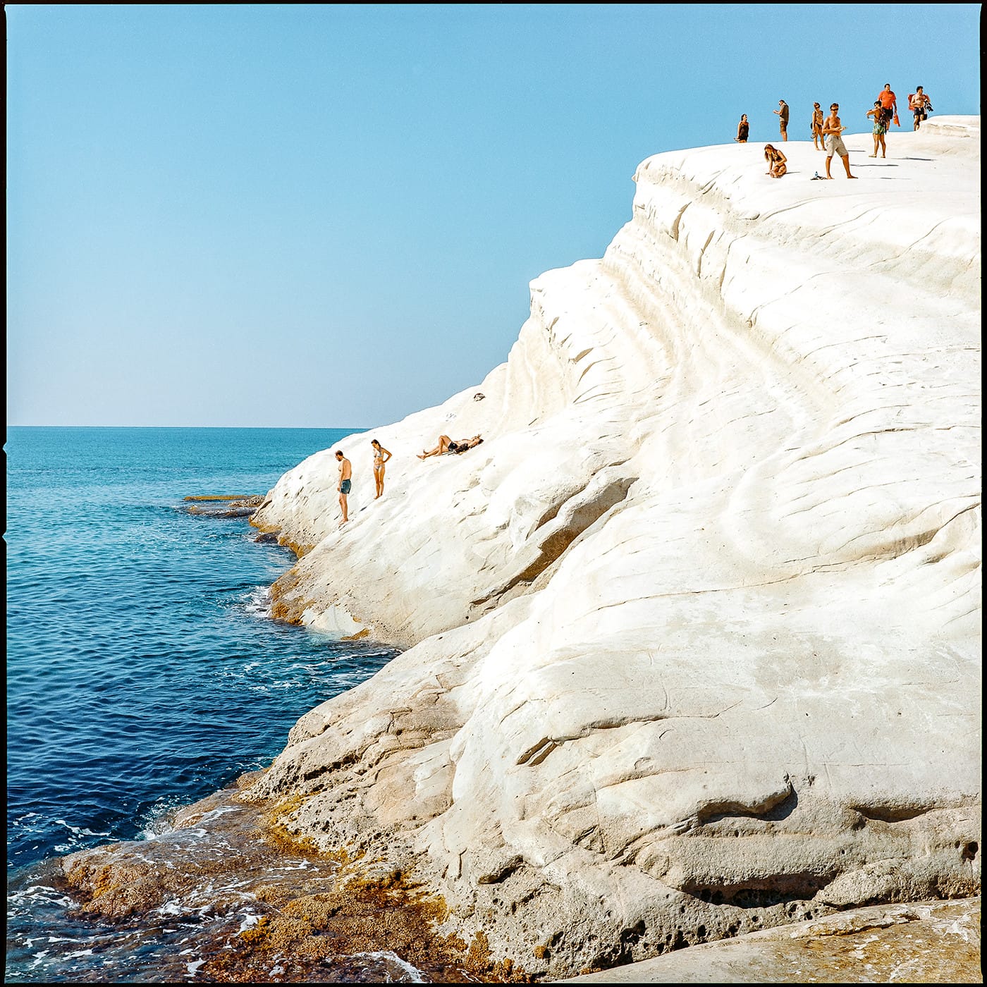 White chalk cliffs with swimmers and sunbathers on terraced limestone formations Sicily.