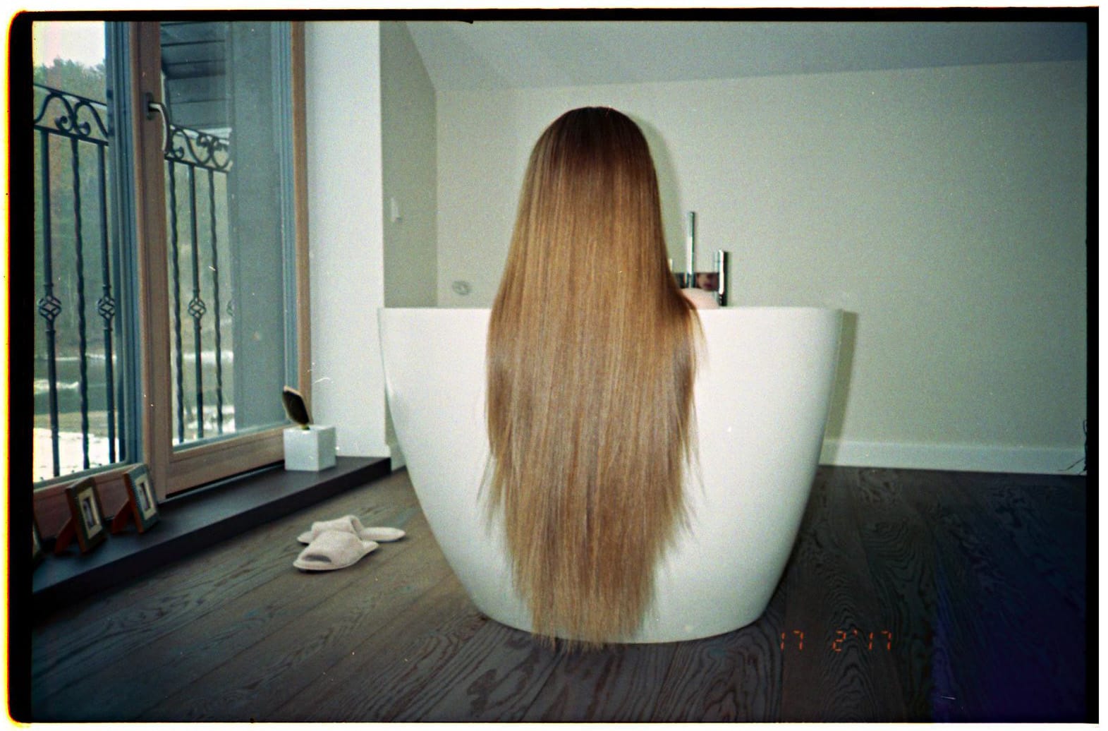 Brunette with long hair sitting in modern white freestanding bathtub near balcony window.