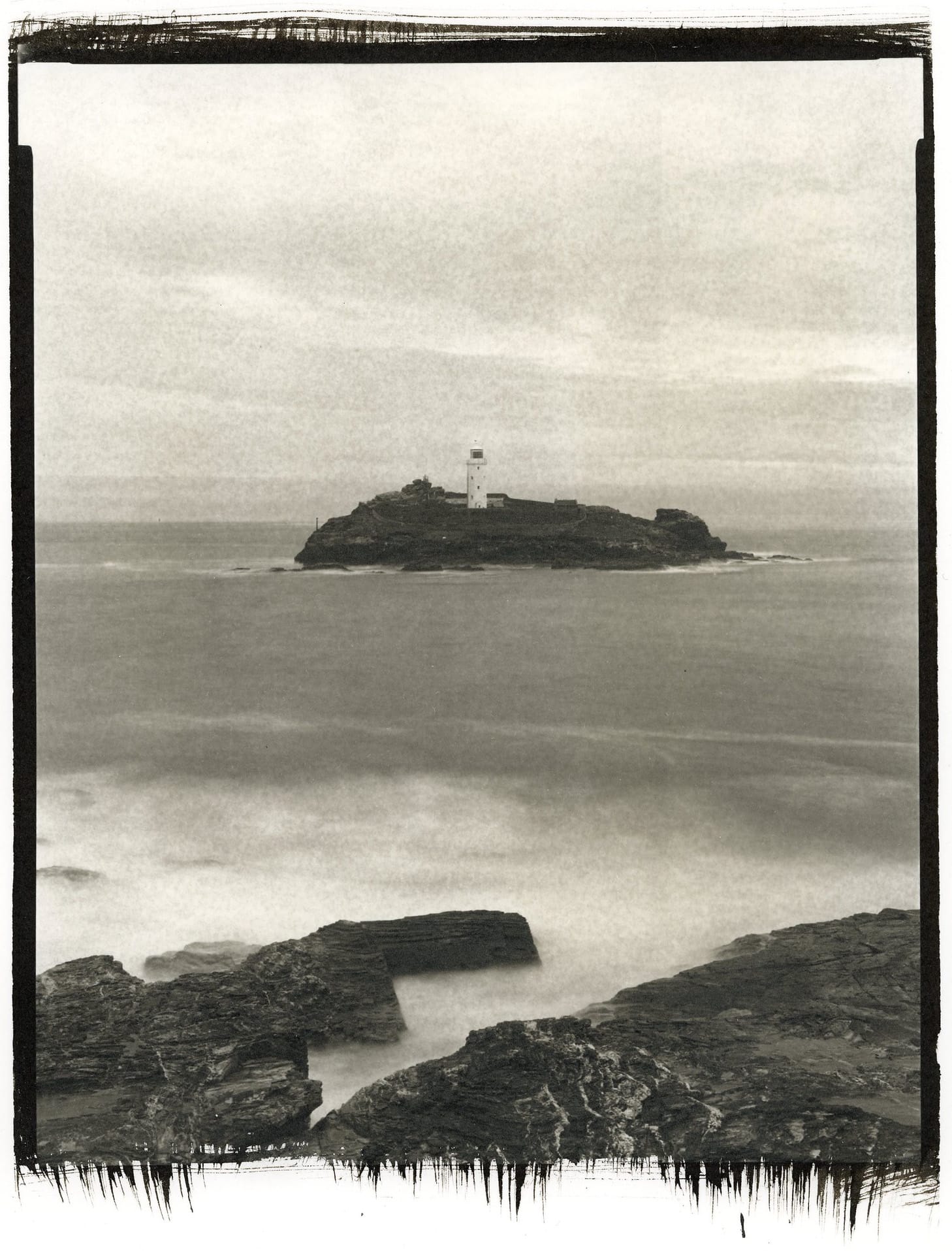 Lighthouse on rocky island surrounded by misty water long exposure black white.