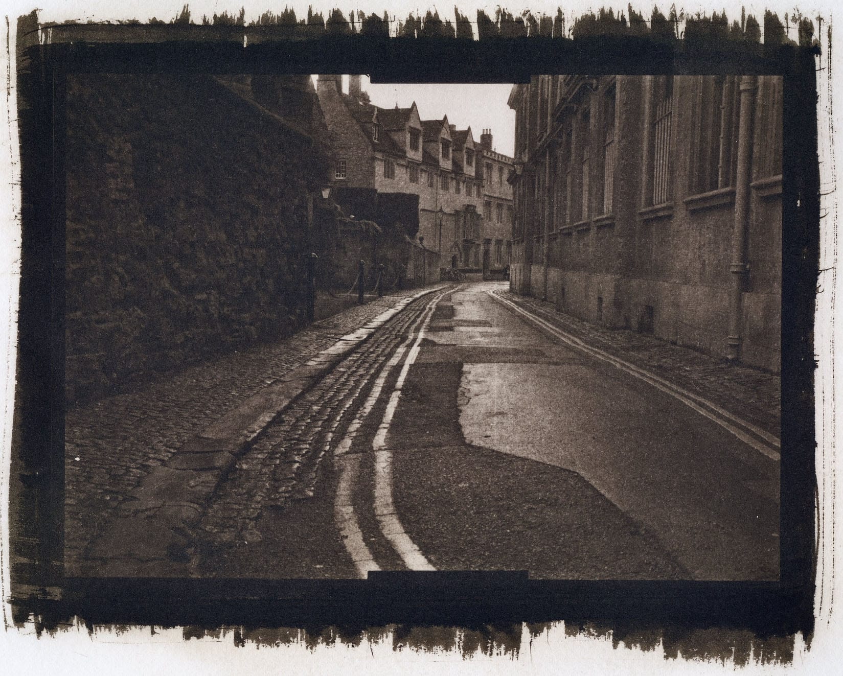 Narrow cobblestone alley under archway with wet pavement and row houses.